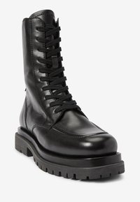 Czarne skórzane buty bojowe z przodem na sznurówki, okrągłym czubkiem, grubą teksturowaną gumową podeszwą i wyraźnymi detalami przeszycia.
