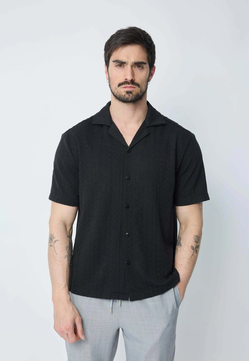 Chemise noire à manches courtes avec un motif texturé, col ouvert et boutons centraux. Associée à un pantalon gris clair. Motifs de tomates sur le tissu.