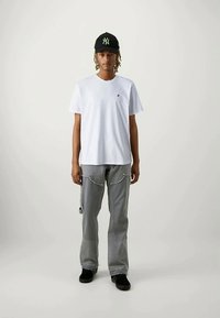 Cilvēks ar gaiši brūniem pīnēm, valkājot melnu cepuri, baltu T-kreklu, pelēkus cargo šortus un melnus sporta apavus, stāv pret vienkāršu fona.