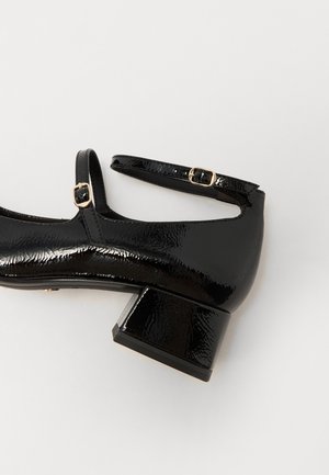 Chaussure Mary Jane en cuir verni noir avec petit talon bloc et deux brides réglables à boucle dorée sur fond blanc.