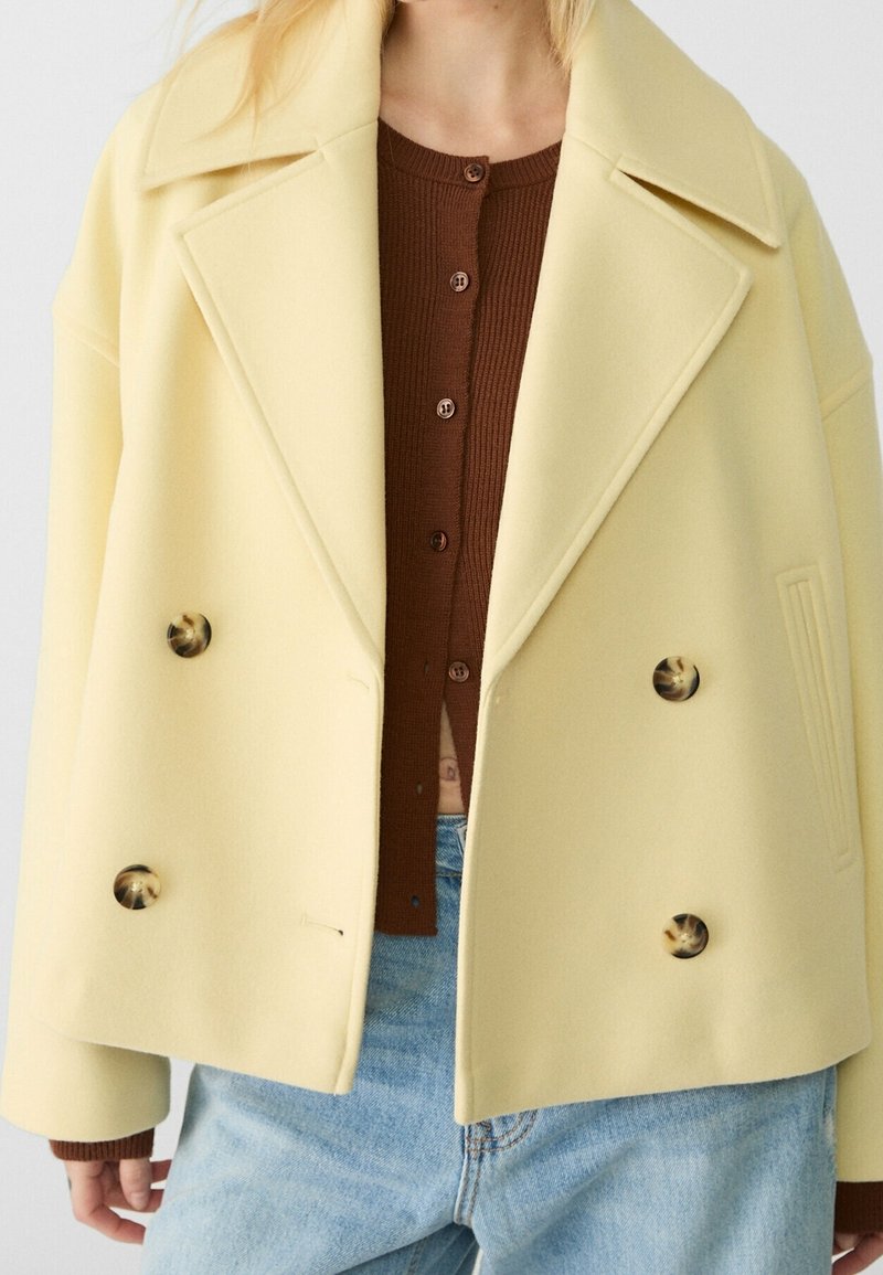 Personne portant un manteau croisé crème sur un pull marron boutonné et un jean bleu clair, debout devant un fond uni.