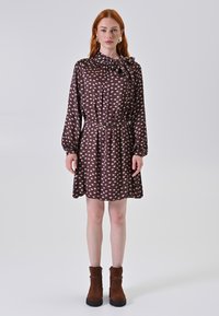 Braun gemustertes Kleid mit Schleifenkragen, langen Ärmeln und einem elastischen Taillenbund. Dazu braune Knöchelstiefel getragen. Glatte Stofftextur.