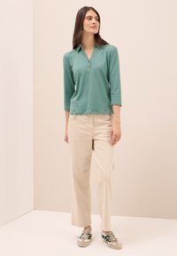 Lange mouwen, kraagloze teal top, gecombineerd met beige wijde broek en grijs-groene sneakers met groene accenten. Gladde stof, relaxte pasvorm.