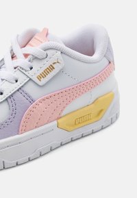 Zapatilla blanca con acentos en rosa y morado, de cuero texturizado y detalle de logo dorado. Presenta una suela blanca gruesa y diseño con cordones.
