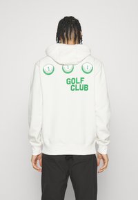 Vit hoodie med en framficka som en kenguru, med grön text "GOLF KLUBB" och tre golfbollar som grafik på baksidan. Slät textur.