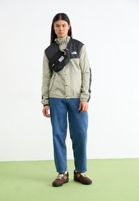 Giacca grigia chiara e nera con bottoni a pressione, abbinata a jeans blu e sneakers marroni. Una piccola borsa a tracolla nera è indossata sopra la giacca.