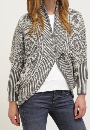 Femme porte un cardigan en maille gris et blanc à motif avec col châle, par-dessus une chemise blanche et un jean foncé.