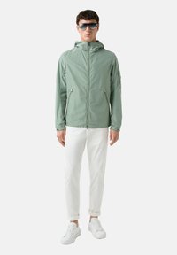Light green lightweight jacket with a hood, zippered front, and side pockets, worn over white rolled-up trousers and white sneakers.