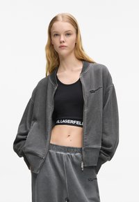 Chaqueta bomber gris con cremallera en tela cepillada, llevada sobre un top corto negro. Presenta mangas anchas y un sutil logo en ambas prendas.