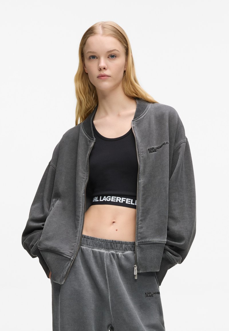 Chaqueta bomber gris con cremallera en tela cepillada, llevada sobre un top corto negro. Presenta mangas anchas y un sutil logo en ambas prendas.