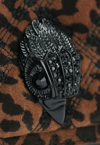 Spilla decorativa nera a forma di uccello, caratterizzata da piume testurizzate e pietre dure nere, su uno sfondo con stampa leopardata.