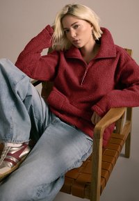 Pulover roșu din fleece cu fermoar până la jumătate și buzunar frontal; asortat cu blugi bleu deschis, lejeri, și adidași argintii cu accente marsala.