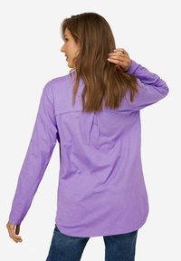 Camicia a maniche lunghe viola con una vestibilità rilassata, dettagli con pieghe sul retro e tessuto morbido e leggero. Abbinata a jeans blu.