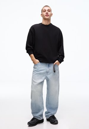 Mladý muž v čiernej mikine, voľných svetlomodrých džínsach s kľúčenkou a čiernych teniskách stojaci s rukami v vreckách.