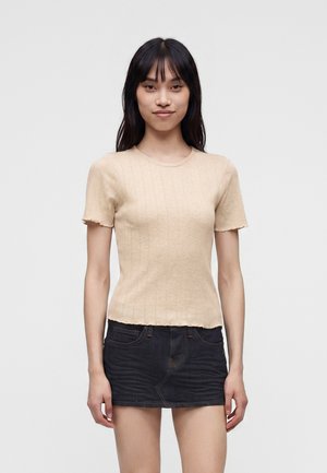Ung kvinde med langt sort hår iført en beige kortærmet top og sort denim mini nederdel, stående mod en ensartet lys baggrund.