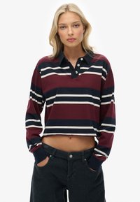 Femme blonde portant un t-shirt rugby court à manches longues avec des rayures bordeaux, marine et blanches, et un pantalon en velours côtelé noir, les mains dans les poches.