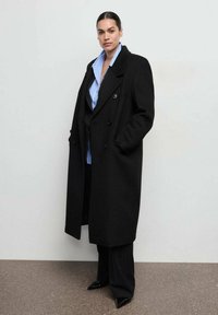Czarny wełniany płaszcz dwurzędowy z szerokimi klapami i bocznymi kieszeniami. Noszony rozpięty na niebieską koszulę z kołnierzykiem i ciemne, dopasowane spodnie.