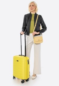 Yellow hard-shell suitcase with black zipper and handle, standing on four wheels, alongside a beige handbag. Model wears a black jacket and green top.