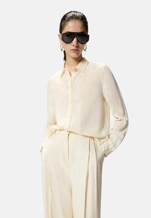 Mujer con camisa cremosa de manga larga abotonada y pantalones de talle alto a juego, con gafas de sol negras de gran tamaño y pendientes de aro dorados.