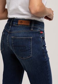 Osoba nosząca ciemnoniebieskie dżinsy Lee Cooper i białą koszulę, z bliska pokazująca tylną kieszeń i okolice talii.