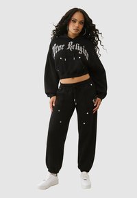 Zwarte cropped hoodie met het logo van "True Religion", gecombineerd met losse zwarte broek met zakken en decoratieve studs. Draagt witte sneakers.