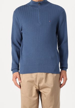 Pull en tricot bleu avec un col haut et une fermeture éclair mi-zip. Motif en chevrons texturé. Poignets et ourlet côtelés. Porté avec un pantalon beige.