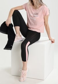 Žena v růžovém tričku a černých legínách s růžovým pruhem sedí na bílém kvádru, drží černý oděv a má na nohou růžové tenisky.