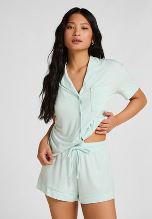 Vrouw met lang donker haar, gekleed in een lichtblauw kort mouwens overhemd met knopen en bijpassende short met trekkoord, poserend tegen een effen achtergrond.