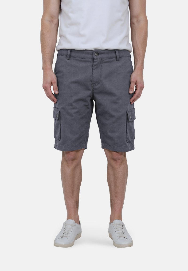 Man in grijze cargoshorts en witte sneakers, staand tegen een effen lichte achtergrond met handen ontspannen naast zich.