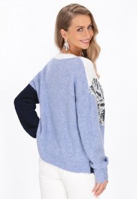 Jasnoniebieski sweter o puszystej fakturze, z kontrastującymi ciemnoniebieskimi rękawami i białym kołnierzykiem. Zawiera dekoracyjny wzór z cekinów z przodu.
