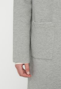 Ανοιχτό γκρι ένδυμα με μακριά μανίκια και υφή υφάσματος, με τσέπη στο μπροστινό μέρος και άσπρες λεπτομέρειες στα μανίκια. Κοντινή λήψη του μανικιού και του χεριού.