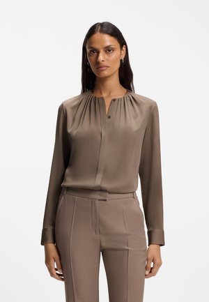 Mujer con cabello largo y oscuro que lleva una blusa de seda color topo con pliegues en el escote y pantalones a medida a juego, sobre un fondo blanco.