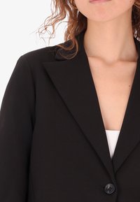 Blazer nero su misura con rever a punta, chiusura a bottone singolo e tessuto liscio; presenta un design aderente senza decorazioni.