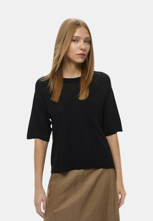 Top corto nero a maniche corte realizzato in un tessuto leggero e liscio, caratterizzato da una piccola tasca sul petto e una vestibilità rilassata. Indossato con una gonna beige.