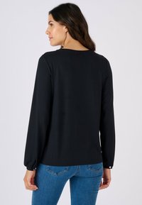 Top nero a maniche lunghe con scollo rotondo e polsini con bottoni, indossato con jeans aderenti blu, che mostra una texture liscia e un design semplice.