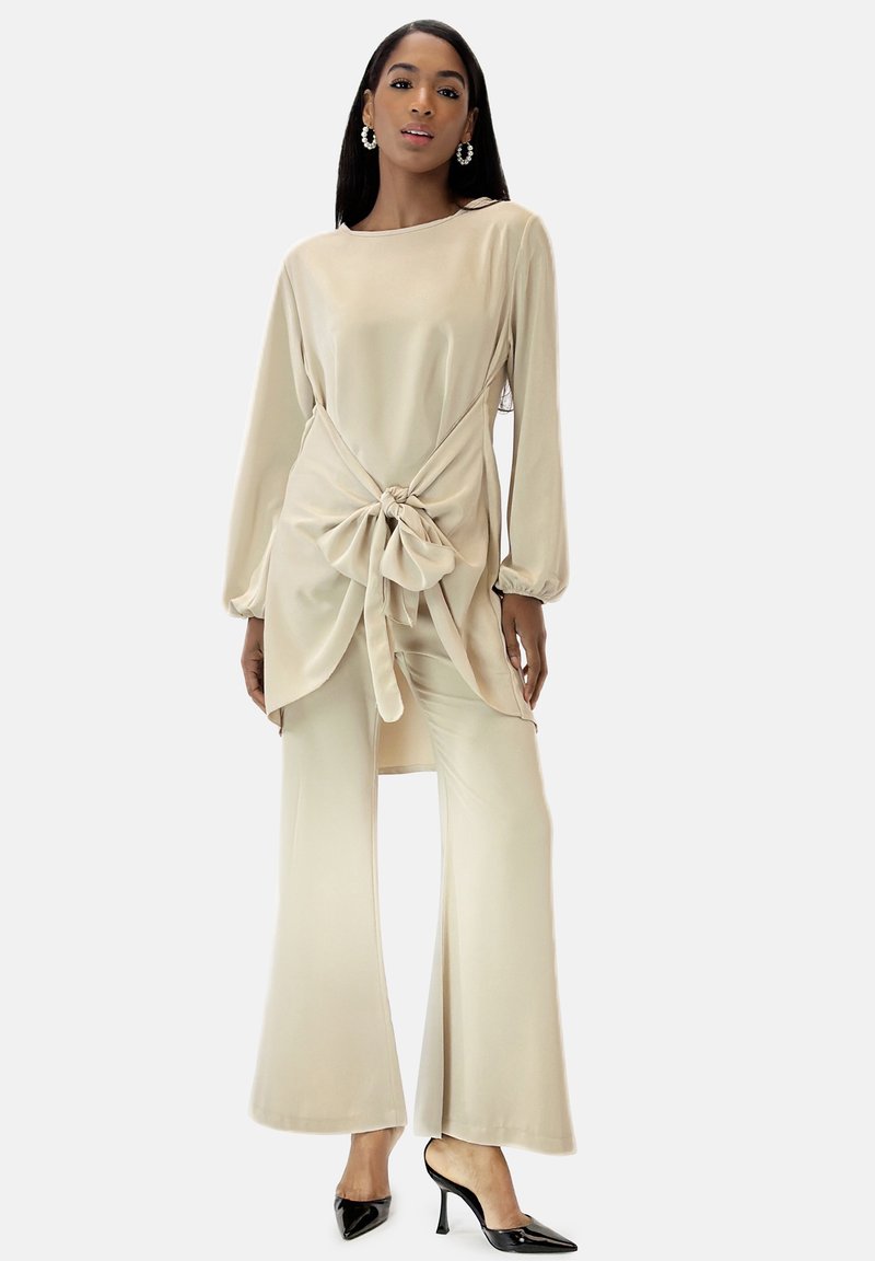 Beige zweiteiliges Outfit mit einem langärmligen Oberteil, das eine vordere Knotendetail aufweist, und weitem Hosenbein. Glatter Stoff mit einem dezenten Glanz.