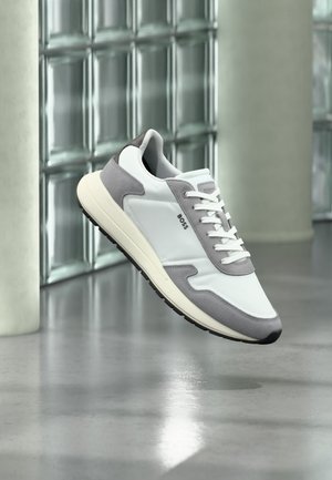 Baskets BOSS blanches et grises avec des lacets blancs flottant au-dessus d'un sol gris réfléchissant, mur en blocs de verre et pilier cylindrique en arrière-plan.