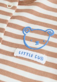 Gros plan sur un tissu à rayures beige et blanc avec une silhouette de visage d'ours bleue et l'inscription brodée "PETIT OURSON" en dessous.