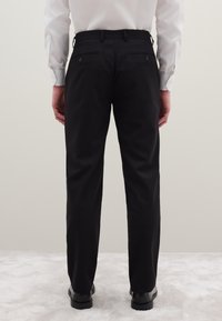 Zwarte broek met een slanke pasvorm, voorzien van twee achterzakken met knopen, gemaakt van een gladde stof, gecombineerd met zwarte schoenen.