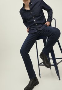 Man som bär en mörk denimskjorta, jeans och svarta stövlar, sitter på en sliten blå metalstol med sidan mot kameran framför en enkel bakgrund.