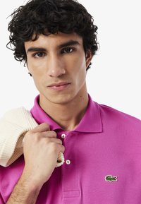 Rosa polo-tröja med två knappar och en liten grön krokodil-logotyp. Modellen håller en krämfärgad stickad tröja över axeln.