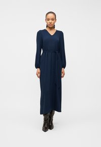 Γυναίκα με πλεξούδες, που φοράει μακρυμάνικο ναυτικό μίντι φόρεμα με ζώνη στη μέση και σκουρόχρωμες άκρες παπούτσια με μυτερά δάχτυλα, στέκεται σε λευκό φόντο.