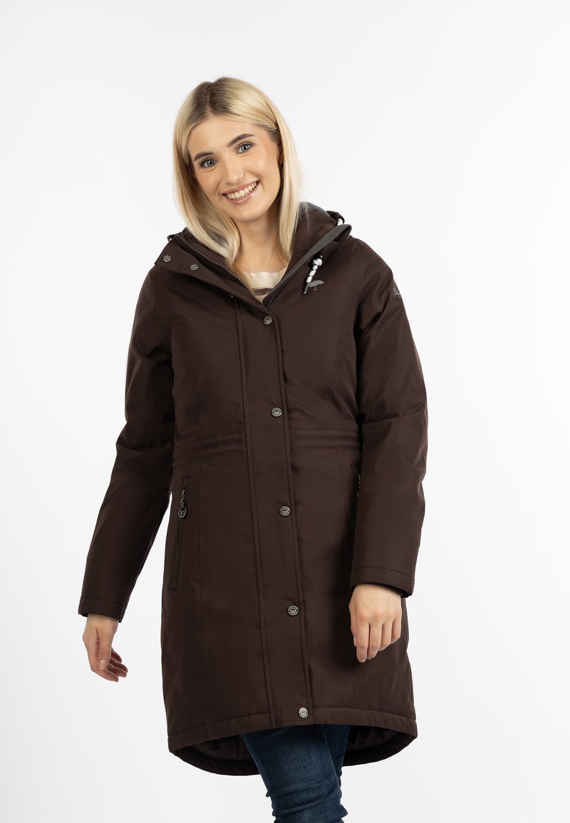 Donna sorridente che indossa un lungo cappotto invernale marrone scuro con cappuccio e tasche con zip, in piedi su uno sfondo bianco.