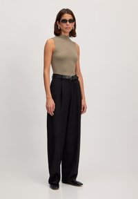 Geribbelde, hoge hals, mouwloze top in olijfgroen, gecombineerd met zwarte, wijde broek en een zwarte riem. Model draagt zwarte zonnebril.
