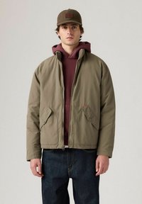 Man i olivgrön zip-up jacka över maroon hoodie och mörk jeans, med en brun rutig keps, står mot en enfärgad bakgrund.
