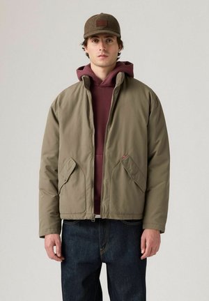 Hombre con chaqueta olive green con cremallera sobre sudadera marón y jeans oscuros, con una gorra a cuadros marrón, de pie contra un fondo liso.