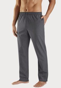 Apsinuogęs vyras, vilkintis juodai baltus smulkiai languotus pižamos kelnes su raišteliu, stovi basomis, viena ranka kišenėje.