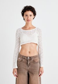 Baltas megztas croptopas su ilgomis rankovėmis, pasižymintis tekstūruotu raštu. Derinamas su rudais džinsais iki liemens su mygtukų uždarymu.