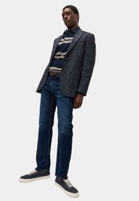 Blazer a quadri grigi sopra una polo a righe blu navy, jeans blu e sneaker navy con suole bianche. Tessuto morbido e vestibilità rilassata.