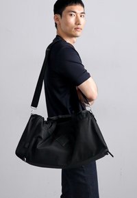 Black duffle bag made of textured synthetic material with a zippered closure and adjustable strap, featuring a diamond-shaped logo accent.